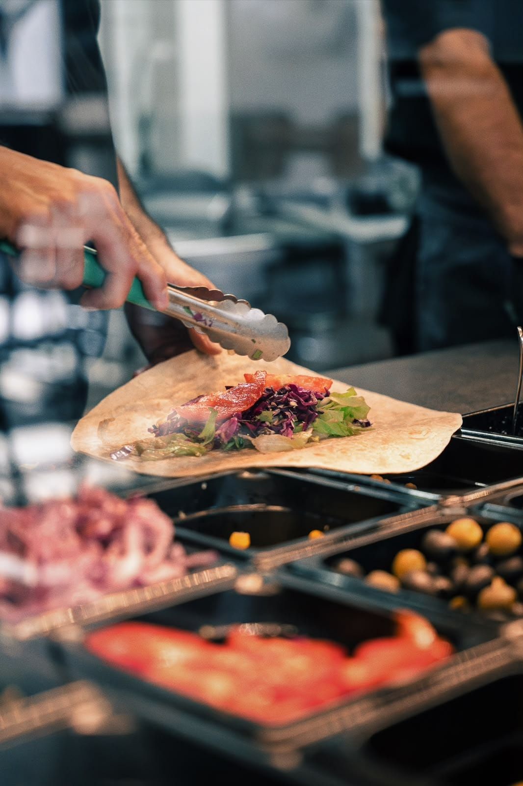 Ein Wrap wird mit der Zange an der Toppings-Theke belegt, davor Schalen mit Tomaten, Oliven, Mais und Rotkraut